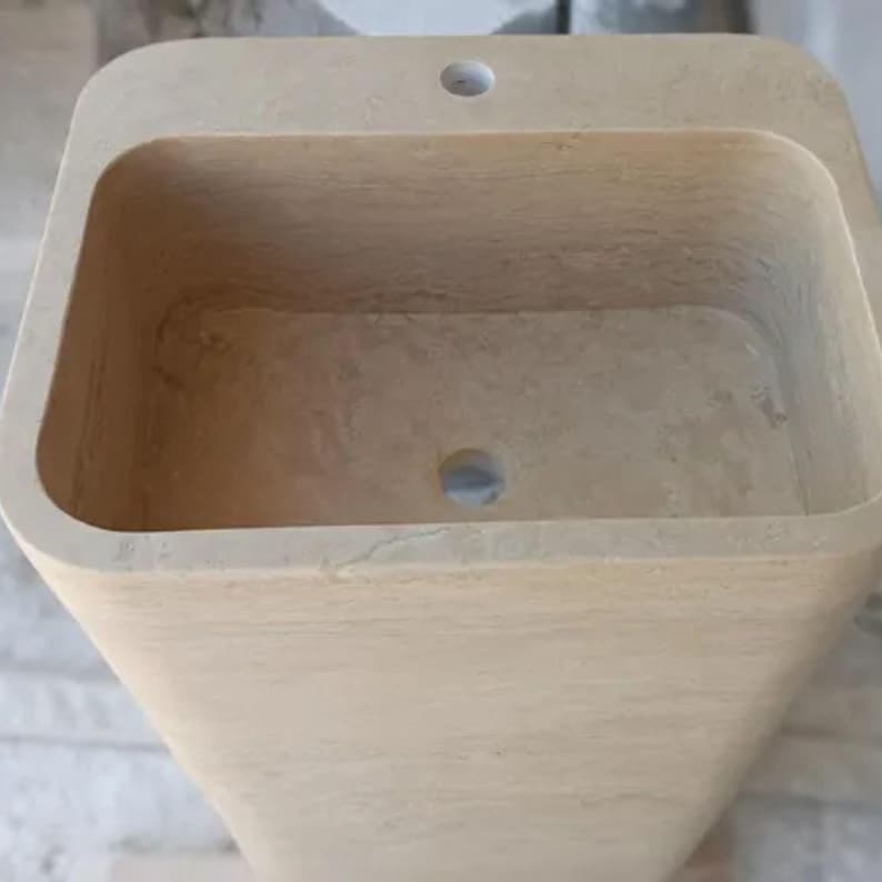 May include: A rectangular beige stone pedestal sink with a smooth, matte finish. The basin features a deep, hollowed-out center with a circular drain hole at the bottom and a single faucet hole centered on the flat top ledge of the basin rim.