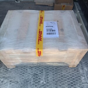 A large, rectangular wooden pallet wrapped in clear plastic. A yellow DHL tape secures the package, with a shipping label attached. The pallet is inside a vehicle.