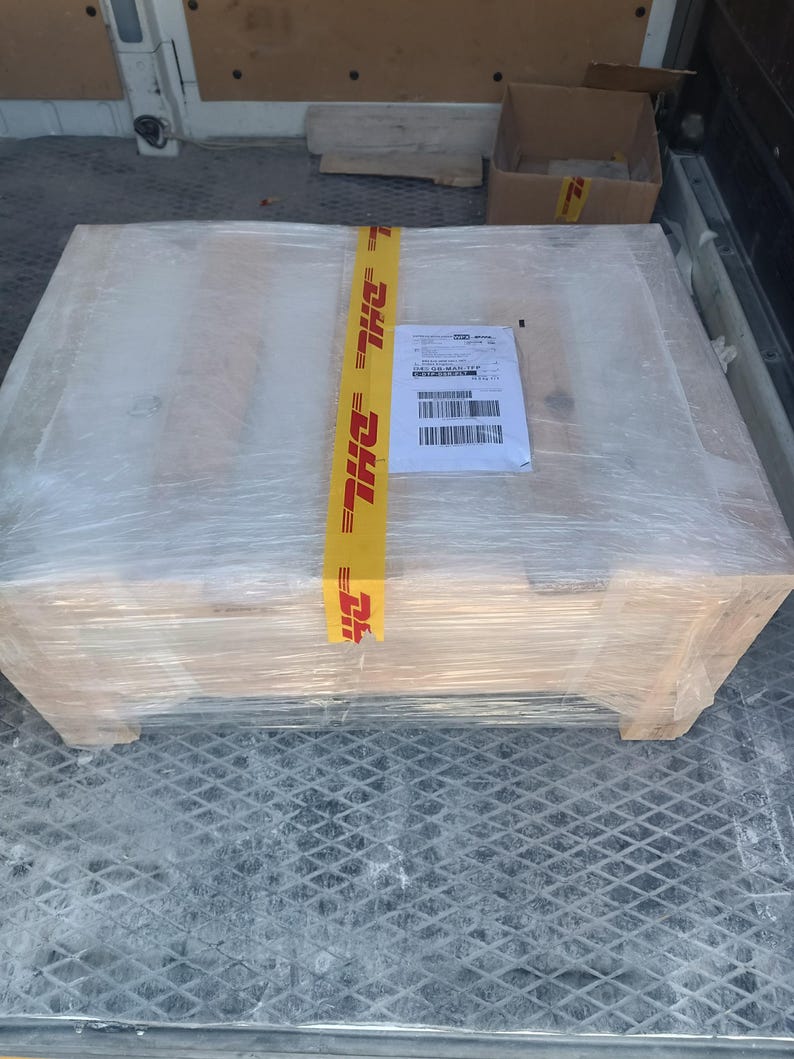 A large, rectangular wooden pallet wrapped in clear plastic. A yellow DHL tape secures the package, with a shipping label attached. The pallet is inside a vehicle.