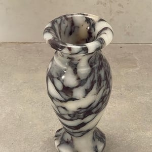 May include: A small, polished stone vase featuring a distinct marbled pattern with swirling veins of deep purple and dark gray set against a creamy white background. The vase has a flared rim, a narrow neck, and a rounded body on a circular base.