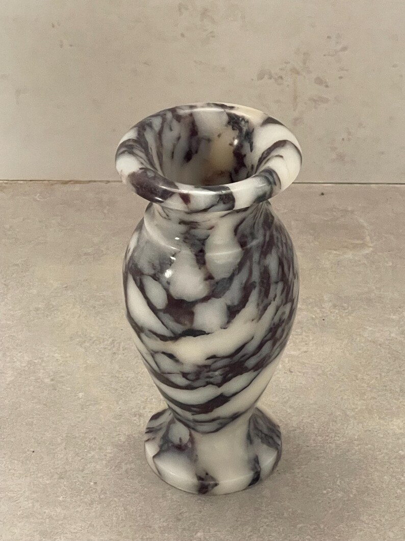 May include: A small, polished stone vase featuring a distinct marbled pattern with swirling veins of deep purple and dark gray set against a creamy white background. The vase has a flared rim, a narrow neck, and a rounded body on a circular base.