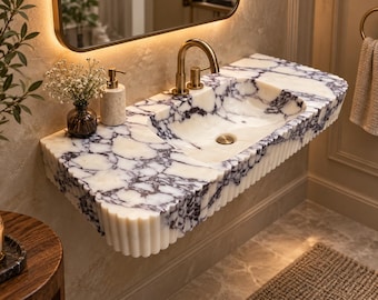 May include: A wall-mounted bathroom vanity sink crafted from white marble with prominent dark purple veining. The basin features a fluted, rounded front edge and is equipped with a gold-toned faucet and a matching circular drain plug.