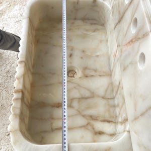 This luxurious Calacatta Gold marble sink features a standard drain hole with a back panel and a striking, elegant design with prominent grey and yellow veins on the front. It can be wall-mounted or countertop-mounted. The front has a fluted design. Dimensions: 24 inches long, 5 inches high, 14 inches deep, panel height 8 inches.