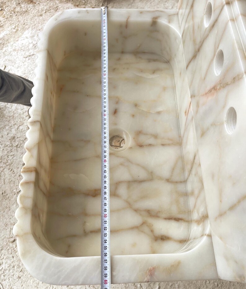 This luxurious Calacatta Gold marble sink features a standard drain hole with a back panel and a striking, elegant design with prominent grey and yellow veins on the front. It can be wall-mounted or countertop-mounted. The front has a fluted design. Dimensions: 24 inches long, 5 inches high, 14 inches deep, panel height 8 inches.