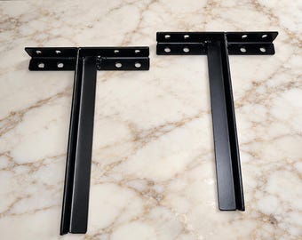 May include: Two black metal T-shaped shelf brackets with a smooth finish. Each bracket features a horizontal top bar with four pre-drilled circular mounting holes and a vertical support arm extending downward, constructed from sturdy steel components.