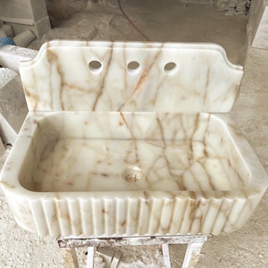 This luxurious Calacatta Gold marble sink with a back panel features a standard drain hole and a striking, elegant design with prominent grey and yellow veins on the front. It can be wall-mounted or countertop-mounted. The front has a fluted design.