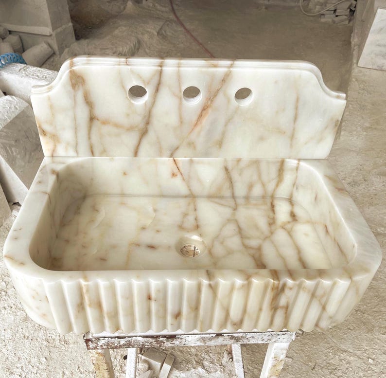 This luxurious Calacatta Gold marble sink with a back panel features a standard drain hole and a striking, elegant design with prominent grey and yellow veins on the front. It can be wall-mounted or countertop-mounted. The front has a fluted design.