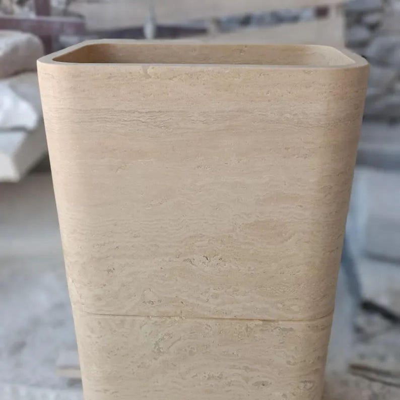 May include: A tall, rectangular vase crafted from light beige natural stone. The surface features a distinct horizontal banded texture with subtle variations in color and porous markings characteristic of the stone material throughout the exterior.