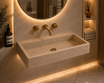 Puede incluir: Un lavabo rectangular montado en la pared, fabricado en piedra beige con textura. La cubeta cuenta con una tapa de desagüe de piedra a juego y se complementa con un grifo de pared de latón, que incluye un caño central y dos mandos laterales.