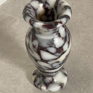 May include: A small stone vase featuring a polished surface with a marbled pattern of white and deep dark brown veins. The vessel has a rounded body, a flared rim, and a circular base, showcasing the natural variations of the stone material.