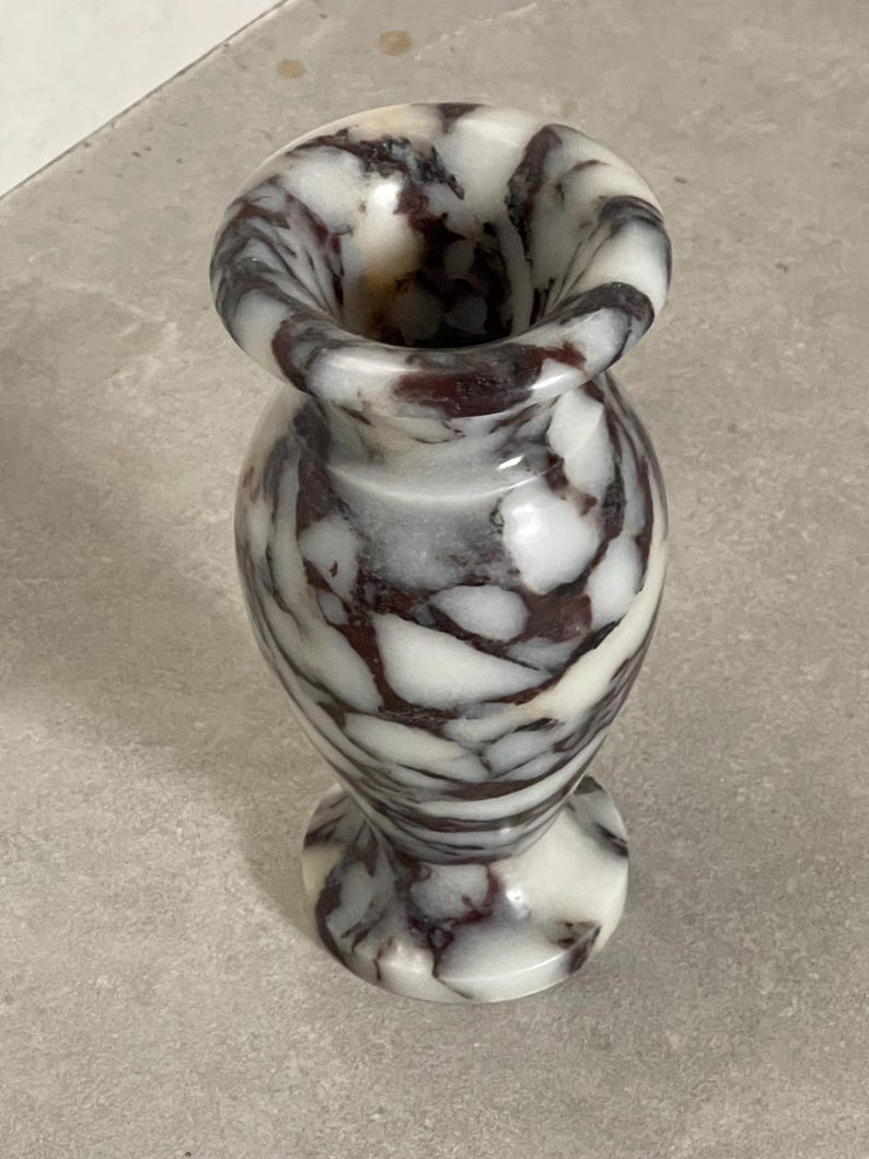 May include: A small stone vase featuring a polished surface with a marbled pattern of white and deep dark brown veins. The vessel has a rounded body, a flared rim, and a circular base, showcasing the natural variations of the stone material.