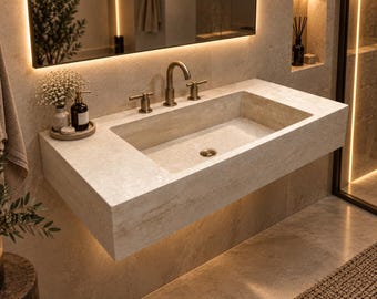 May include: A wall-mounted rectangular bathroom sink crafted from beige natural stone with a textured finish. It features a central basin, a brass faucet with two matching side handles, and a circular brass drain cover centered in the basin floor.