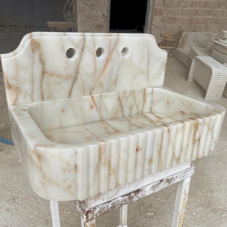 This luxurious Calacatta Gold marble sink with a back panel features a standard drain hole and a striking, elegant design with prominent grey and yellow veins on the front. It can be wall-mounted or countertop-mounted. The front has a fluted design.