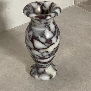 May include: A small stone vase featuring a polished surface with a marbled pattern of white, grey, and deep burgundy veins. The vessel has a flared rim, a narrow neck, a rounded body, and a circular base, showcasing natural stone variations.
