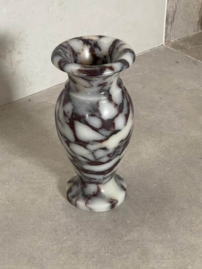 May include: A small stone vase featuring a polished surface with a marbled pattern of white, grey, and deep burgundy veins. The vessel has a flared rim, a narrow neck, a rounded body, and a circular base, showcasing natural stone variations.