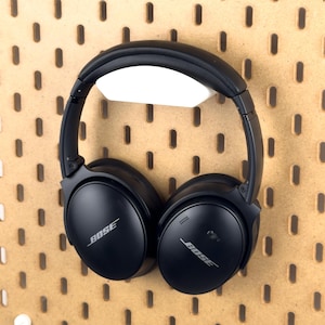 Könnte beinhalten: Schwarzer Bose-Kopfhörer, der an einer hellbraunen Lochwand hängt. Die Lochwand hat gleichmäßig verteilte Löcher und weiße Befestigungselemente. Der Kopfhörer wird von einem weißen Halter gehalten.