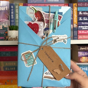 May include: A light blue wrapped gift tied with twine, featuring a brown tag that reads "romance" with a butterfly cutout. The gift is decorated with stickers and contains a red heart, a pen, and other small items. Bookshelves are in the background.