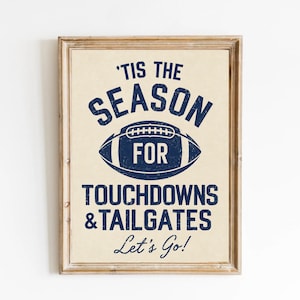 Könnte beinhalten: Ein gerahmter Kunstdruck mit cremefarbenem Hintergrund und dunkelblauem, abgenutztem Text, der 'TIS THE SEASON FOR TOUCHDOWNS & TAILGATES Let's Go!' lautet, zentriert um eine grafische Abbildung eines American Footballs in der Mitte.