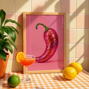 May include: A framed art print featuring a red disco ball chili pepper on a pink background. The chili is covered in small reflective mirror tiles. The frame is light wood, and the print is displayed on an orange and white checkered surface.