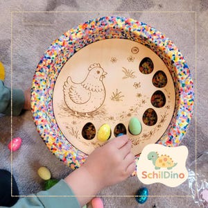 Könnte beinhalten: Osterei-Sortierspiel aus Holz mit farbenfrohem Rand. In der Mitte befindet sich ein lasergraviertes Hühner- und Grasdesign. Enthält sechs bunte Eier. Eine Kinderhand greift nach einem Ei. Das Logo "SchildDino" befindet sich unten rechts.