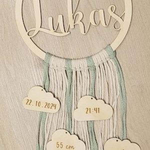 Könnte beinhalten: Holz-Kinderzimmerdekoration mit dem Namen "Lukas" in kursivschriftlicher Schrift auf einem runden Holzreifen. Der Reifen ist mit einer Makramee-Fransenborte und vier Holz-Wolken-Etiketten mit dem Text "22.10.2024", "21:41", "55 cm" und "3.650 g" verziert.