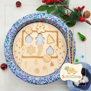 Könnte beinhalten: Ein Holzpuzzle mit Weihnachtsmotiven, darunter Lebkuchenmännchen und Tannenbaum. Das Puzzle befindet sich in einer blau-violett gesprenkelten Schüssel. Dekorative Elemente sind rote Ornamente und Grünzeug.