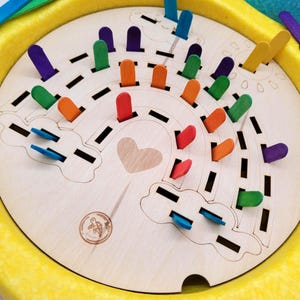 Könnte beinhalten: Holz-Regenbogen-Puzzle mit bunten Eisstielen. Das Puzzle zeigt eine Herzform und Wolkenumrisse. Die Eisstiele sind in verschiedenen Farben gehalten, darunter Rot, Orange, Gelb, Grün, Blau und Lila. Das Puzzle befindet sich in einer gelben Schale.