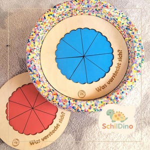 Könnte beinhalten: Pädagogisches Holzspielzeug mit einem Design aus blauen und roten Segmentkreisen. Das Spielzeug hat einen bunten, gesprenkelten Rand und der Text "Was versteckt sich?" ist auf die Holzoberfläche gedruckt. Das Spielzeug ist für Kinder konzipiert.