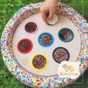 Könnte beinhalten: Ein sensorisches Holzspielzeug mit bunten, runden Einsätzen. Das Spielzeug hat einen mehrfarbigen Stoffrand und eine Kinderhand legt eine Holzscheibe auf die Oberfläche. Das Spielzeug hat das Logo "Schildino" unten rechts.