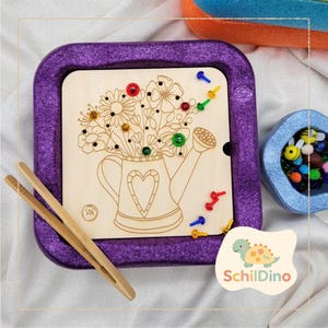 Könnte beinhalten: Holz-Steckbrettspiel mit Blumenmotiv und lila Rahmen. Das Brett zeigt ein Gießkannen-Design mit Herz und Blumen. Inklusive farbigen Stiften und Holzpinzette. Ein kleiner Behälter mit bienenförmigen Perlen ist ebenfalls sichtbar.