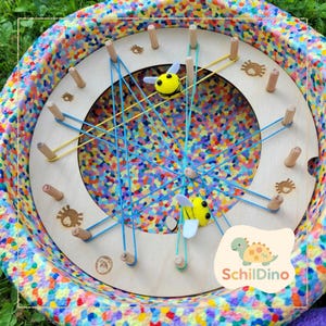 Könnte beinhalten: Ein pädagogisches Holzspielzeug mit kreisförmigem Design, das Stifte, bunte Gummibänder und bienenförmige Dekorationen aufweist. Das Spielzeug befindet sich in einem mehrfarbigen, gesprenkelten Behälter. Das Logo "Schildino" ist unten rechts sichtbar.