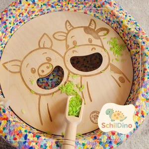 Könnte beinhalten: Sensorisches Holzspielzeug mit Schwein- und Kuhdesign, gefüllt mit bunten Perlen. Das Spielzeug enthält einen Holzschöpfer und ist von einem bunten, gesprenkelten Rand umgeben. Das Logo "SchildDino" ist unten rechts sichtbar.