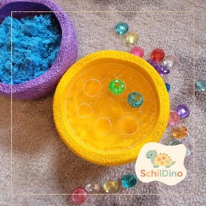 Könnte beinhalten: Eine gelbe sensorische Schale mit sechs kreisförmigen Vertiefungen und bunten Edelsteinen. Eine lila Schüssel mit blauem Sand befindet sich neben der Schale. Das Bild enthält auch das SchilDino-Logo.