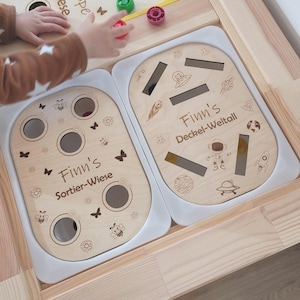 Könnte beinhalten: Ein Holzspielzeug mit zwei separaten Spielbereichen. Die linke Seite hat ein Sortierspiel mit Löchern und dem Text "Finn's Sortier-Wiese". Die rechte Seite hat ein Spiel mit Deckel und dem Text "Finn's Deckel-Weltall".