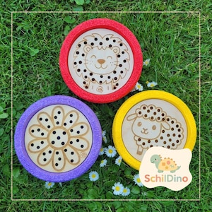 Könnte beinhalten: Drei Holzspielzeuge mit farbigen Rändern, die einen Löwen, eine Blume und ein Schaf zeigen. Die Spielzeuge liegen auf einer grünen Wiese mit kleinen weißen Blumen. Das Markenlogo "SchildDino" ist sichtbar.