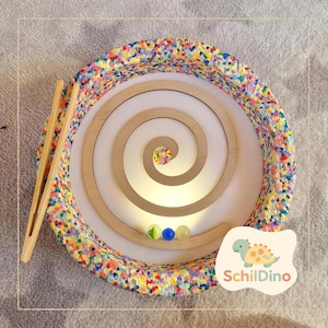 Könnte beinhalten: Ein Holz-Spiral-Labyrinth mit drei Murmeln und einer Holzpinzette. Das Labyrinth ist von einem bunten, gesprenkelten Rand umgeben. Das Bild enthält den Text "Schildino" und einen Cartoon-Dinosaurier.