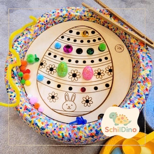Könnte beinhalten: Osterei-Aktivitätstafel aus Holz mit Hasen- und Blumenmuster, umgeben von einem farbenfrohen Rand. Enthält bunte Pompons, Glitzereier und Holzpinzetten zur Förderung der Feinmotorik. Das Brett trägt den Text "SchildDino".