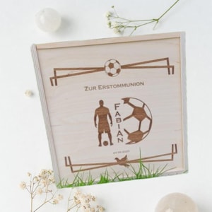 Könnte beinhalten: Eine hellfarbene Holzkiste mit Fußballmotiv. Die Kiste zeigt einen Fußball und eine Silhouette eines Fußballspielers. Der Text "ZUR ERSTKOMMUNION" und der Name "FABIAN" sind ebenfalls sichtbar.