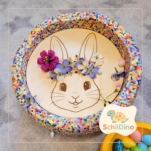 Könnte beinhalten: Ein runder Holztablett mit einem bunten, gesprenkelten Rand. Das Tablett zeigt ein lasergeätztes Hasengesicht mit einem Blumenkranz. Der Hase ist mit lila, blauen und weißen Blumen geschmückt. Das Tablett ist mit kleinen, bunten Pompons und Blumen verziert.