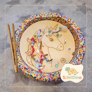 Könnte beinhalten: Ein Holztablett mit einem bunten, gesprenkelten Rand und einer Holzbasis mit einem Einhorn-Design. Das Tablett enthält Perlen, Pfeifenreiniger und kleine Edelsteine. Eine Holzpinzette liegt daneben. Das Logo "SchildDino" befindet sich unten rechts.
