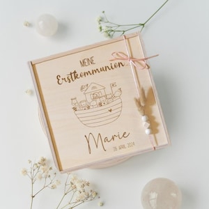 Könnte beinhalten: Eine Holzkiste mit einem hellrosa Band und einem kleinen Strauß weißer Blumen. Die Kiste ist mit dem Text "Meine Erstkommunion" und dem Namen "Marie" mit dem Datum "28. April 2024" graviert. Die Kiste zeigt ein Design eines Bootes mit Tieren darauf.