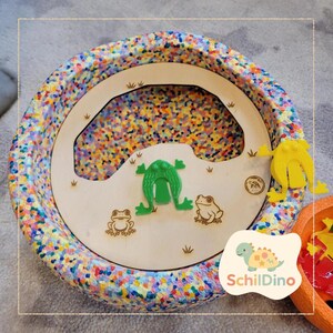 Könnte beinhalten: Ein farbenfrohes sensorisches Spielzeug mit einem Holzmittelteil mit Froschmotiven und einer grünen Froschfigur. Der Rand ist mit verschiedenen Farben gesprenkelt. Das Spielzeug enthält den Text "Schildino" und ist für Kinder konzipiert.