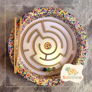 Könnte beinhalten: Ein kreisförmiges Holz-Labyrinth-Spiel mit farbigem Rand und weißem Innenraum. Das Labyrinth hat ein Wegmuster. Drei Murmeln befinden sich unten. Holzstäbchen sind enthalten. Das Logo "Schildino" ist sichtbar.