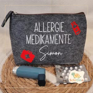 Könnte beinhalten: Eine graue Filz-Reißverschlusstasche mit den Worten "ALLERGIE MEDIKAMENTE Simon" in Weiß. Rote medizinische Symbole und ein Inhalator, eine Blisterpackung mit Pillen und ein kleiner Aufkleber sind sichtbar.