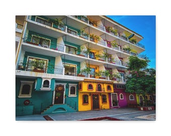 May include: A vibrant, multi-story building with colorful facades. The building features balconies with plants, and the lower levels are painted in shades of teal, yellow, and purple. The sky is a clear blue.