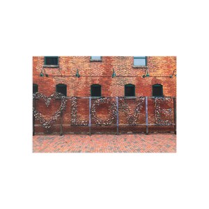 May include: A brick wall with the word "LOVE" spelled out in metal frames. The "O" is a heart shape. The letters are filled with small, colorful locks. The background is a red brick wall with windows.