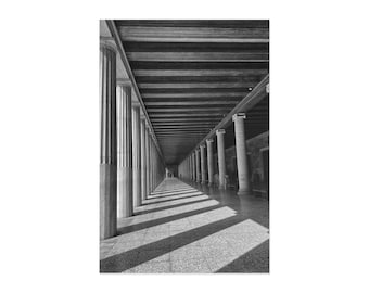 May include: Black and white photograph of a colonnade with a long perspective. The columns are fluted and support a wooden ceiling. Shadows from the ceiling create stripes across the tiled floor. The image is a study in lines and light.