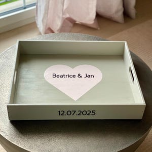Könnte beinhalten: Ein weißes rechteckiges Serviertablett mit Griffen. Das Tablett hat einen hellgrünen Innenraum mit einem rosa Herzdesign in der Mitte, auf dem die Namen "Beatrice & Jan" stehen. Das Datum "12.07.2025" ist darunter aufgedruckt.