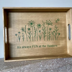 Könnte beinhalten: Serviertablett aus Holz mit hellbraunem Finish. Das Tablett zeigt ein grünes Blumendesign und den Text "It's always FUN at the Bidders". Es hat ausgeschnittene Griffe an den Seiten.