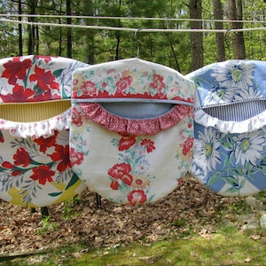 May include: Three fabric bag holders with metal hangers, each featuring a distinct floral pattern. The left bag has red flowers and yellow accents, the center has red floral prints with a ruffled edge, and the right has blue and white daisies.